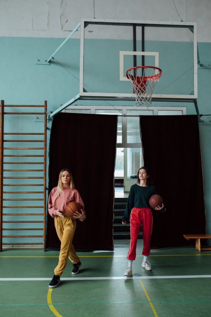 Two women in trendy sportswear holding basketballs indoors.